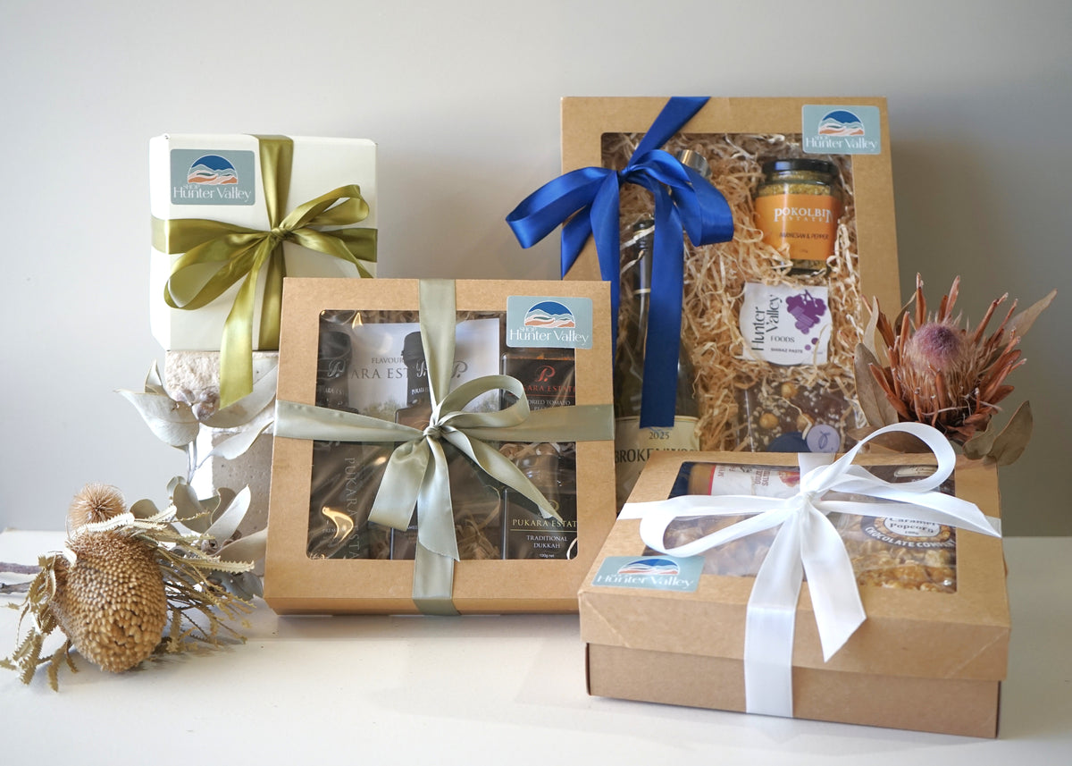 Three gift boxes from The Gift Shop arranged on a white surface against a light gray wall. The boxes are wrapped in brown kraft paper with satin ribbons in olive green, blue, and white, and feature clear windows showing packaged treats and a jar.